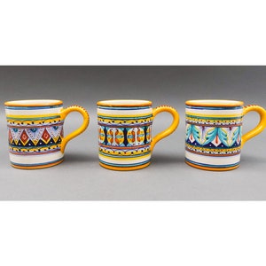 May include: Three ceramic mugs with yellow rims and colourful geometric patterns. The mugs have white interiors and are decorated with blue, red, green, and yellow accents.