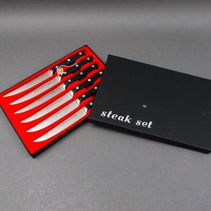 May include: A set of six steak knives with black handles in a red and black box. The box has the words "steak set" printed on it.