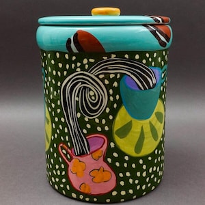Op de afbeelding: Een groene keramische koekjespot met een teal deksel en een gele knop. De pot is versierd met een grillig ontwerp van een roze en oranje vaas met een zwart-witte swirl die eruit komt, en een groene en blauwe mok met een zwart-witte swirl die eruit komt. De achtergrond is groen met witte stippen.