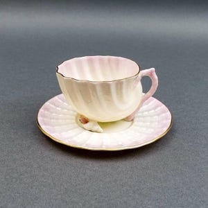 Puede incluir: Un delicado juego de taza y platillo de té en tonos rosa pálido y crema. La taza presenta un diseño en espiral y un borde dorado, con un platillo a juego. El conjunto está acentuado con detalles de conchas marinas, creando una estética costera.