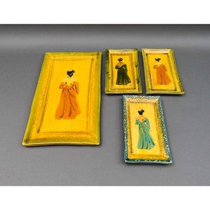 Könnte beinhalten: Set aus fünf rechteckigen Keramiktabletts. Jedes Tablett hat einen gelben Hintergrund und eine gemalte Figur in einem Kleid. Die Kleider sind in Orange, Grün und Türkis gehalten. Die Tabletts haben einen strukturierten, gelben Rand.
