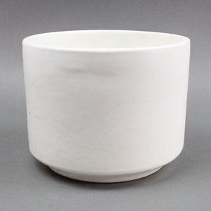 May include: A white ceramic bowl with a cylindrical shape and a slightly flared base. The bowl has a smooth, matte finish and a simple, minimalist design. The interior is empty, and the bowl is set against a gray background.
