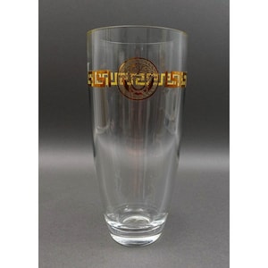 Puede incluir: Un vaso transparente con una banda decorativa dorada y marrón que presenta una cabeza de Medusa y patrones geométricos. El vaso tiene una forma alta y delgada y una base redondeada. El diseño está centrado.