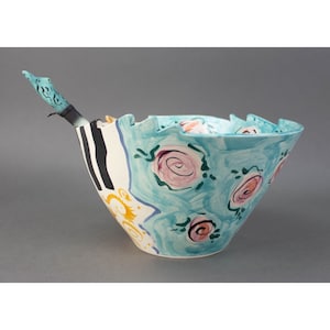 May include: A blue and white ceramic bowl with a whimsical design featuring stylised flowers and a black and white striped pattern. The bowl has a unique, irregular shape and a small, blue bird-shaped handle.