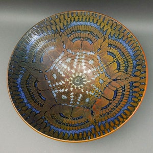 Könnte beinhalten: Eine große Keramikschale mit einer kupfer- und blauen Glasur. Die Schale hat ein kreisförmiges Design mit einem zentralen Motiv aus einem weißen und goldenen Stern.