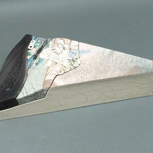 May include: A unique, abstract sculpture with a triangular shape. The sculpture features a black, textured section on one end, transitioning to a silver-toned, cracked surface with blue and white accents. The base is a light gray.