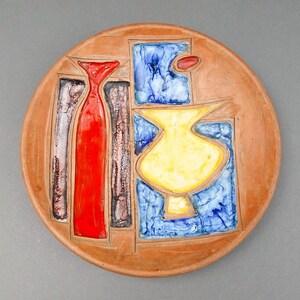 Puede incluir: Un plato redondo de terracota con un diseño estilizado que presenta un jarrón amarillo, rayas verticales rojas y marrones y formas geométricas azules y blancas.
