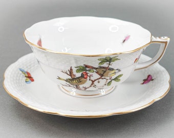 May include: A white porcelain teacup and saucer set with a gold rim. The cup and saucer feature a delicate floral design with two birds perched on a branch. The birds are painted in shades of red, yellow, and green.
