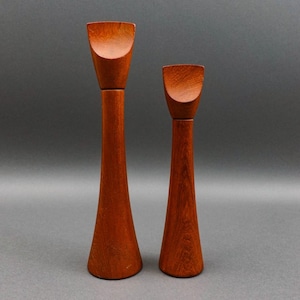 Może przedstawiać: Dwa drewniane świeczniki o ciepłym, czerwonobrązowym wykończeniu. Świeczniki mają unikalny design, z rozszerzoną podstawą i zakrzywioną górą. Wyższy świecznik ma około 25 cm wysokości, a krótszy około 20 cm.