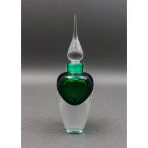 Può includere: Un flacone di profumo in vetro trasparente con un serbatoio verde a forma di cuore e una base smerigliata. Il flacone ha un tappo trasparente e allungato. La parte verde è riempita con un liquido verde scuro. Il flacone è alto circa 15 cm.