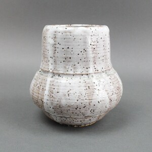 Peut inclure: Un vase en céramique blanche avec une surface texturée et des taches brunes. Le vase a un corps arrondi et une ouverture évasée.