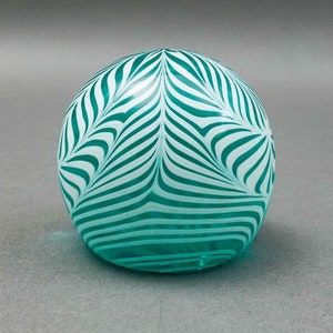 Può includere: Un fermacarte sferico in vetro, turchese e bianco. Presenta un motivo di strisce bianche curve su sfondo turchese, creando un design a vortice. L'oggetto è liscio e riflettente, con una base trasparente.