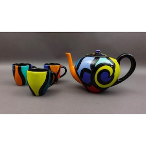 Könnte beinhalten: Ein farbenfrohes Keramik-Teeservice mit einer Teekanne und drei passenden Tassen. Die Teekanne hat einen schwarzen Griff und Deckel mit einem lebendigen Design aus gelben, blauen, roten und schwarzen Wirbeln. Die Tassen haben ein ähnliches Design mit schwarzen Griffen und bunten Mustern.
