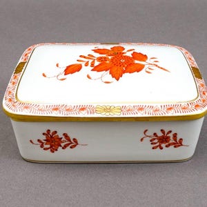 Puede incluir: Una caja cuadrada de porcelana blanca con tapa, decorada con diseños florales naranjas y bordes dorados. La caja mide aproximadamente 12,5 cm de largo.
