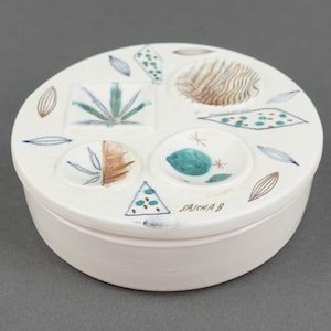 May include: White ceramic round box with a lid. The lid has a geometric design with brown, blue, and green accents. The box is signed "Saicha B".