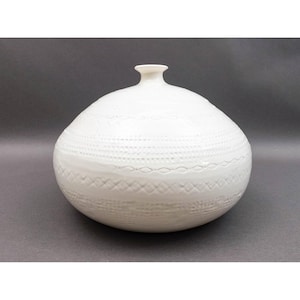 May include: A white ceramic vase with a rounded, bulbous body and a narrow neck. The surface features horizontal bands of embossed geometric patterns, including rows of small dots, diamonds, and cross-hatched textures encircling the center.