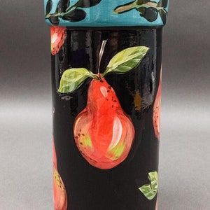 Puede incluir: Un tarro de cerámica negro con una tapa azul y un pomo blanco con lunares. El tarro está decorado con un patrón repetido de peras rojas y hojas verdes.