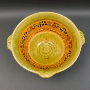 Pode incluir: Uma tigela de cerâmica feita à mão com exterior verde claro e interior amarelo. A tigela apresenta uma faixa decorativa com desenhos florais e de bagas em vermelho, preto e castanho. Possui duas alças e um bico.