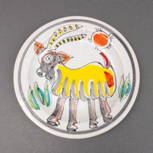 Puede incluir: Un plato de cerámica blanco con un diseño pintado a mano de un animal amarillo y gris con una mancha roja en la espalda. El animal está rodeado de hojas verdes y un sol rojo.