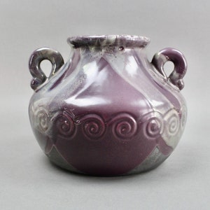 Op de afbeelding: Een paarse keramische vaas met twee handvatten en een decoratief swirl-patroon rond het midden. De vaas heeft een glanzende afwerking en een unieke glazuur.
