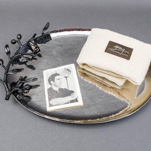 Puede incluir: Una bandeja de plata con acabado martillado presenta un diseño de rama de olivo de metal negro. La bandeja está decorada con una servilleta de tela blanca con el texto "Berg Michael Aram" impreso en ella.