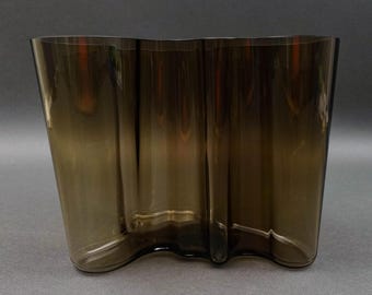 May include: A brown glass vase with a wavy, organic shape. The vase has a translucent quality, allowing light to pass through. The design features a series of vertical, undulating curves, creating a unique and modern aesthetic. The vase is approximately 10 inches tall.