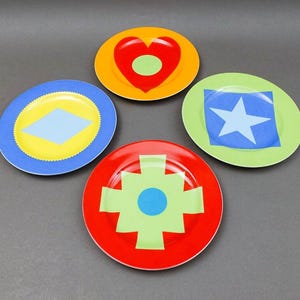 May include: Four decorative plates with colorful geometric designs. Each plate features a different pattern: a heart, a star, a diamond, and a cross-like shape. The plates are in shades of red, yellow, blue, and green.