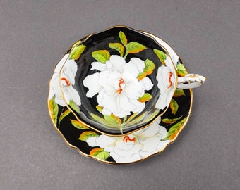 May include: A black and white teacup and saucer set with a floral design. The cup and saucer feature a large white flower with green leaves on a black background.