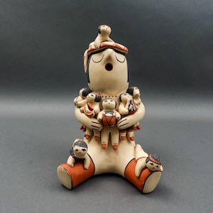 May include: A handcrafted ceramic figurine of a seated figure with a serene expression, adorned with multiple small figures. The main figure is cream-colored with red accents on the legs and headwear. The small figures are held in the arms and seated around the base.
