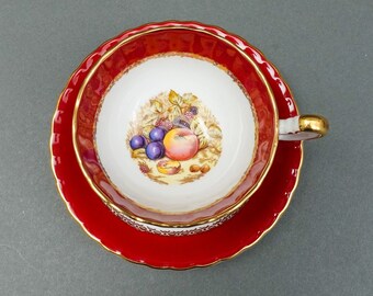 May include: A red teacup and saucer set with a gold rim and a floral design featuring fruit and berries. The saucer has a scalloped edge.