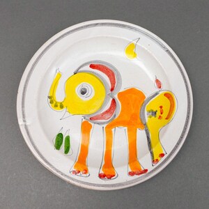 May include: A white ceramic plate with a hand-painted design of an orange and yellow elephant with red and green accents.