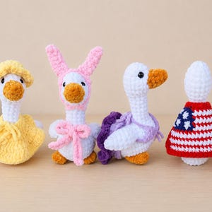 May include: Four crocheted goose figures made of white yarn with orange beaks. The first wears a yellow hat and dress, the second a pink bunny-ear hat and bow, the third a purple shawl, and the fourth a red, white, and blue flag-patterned cape.