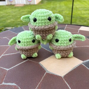 May include: Three handmade crochet figures of a character with large ears and black eyes. The figures are green with a brown base and are arranged on a brown and tan tiled surface. The figures are approximately 10 cm tall.