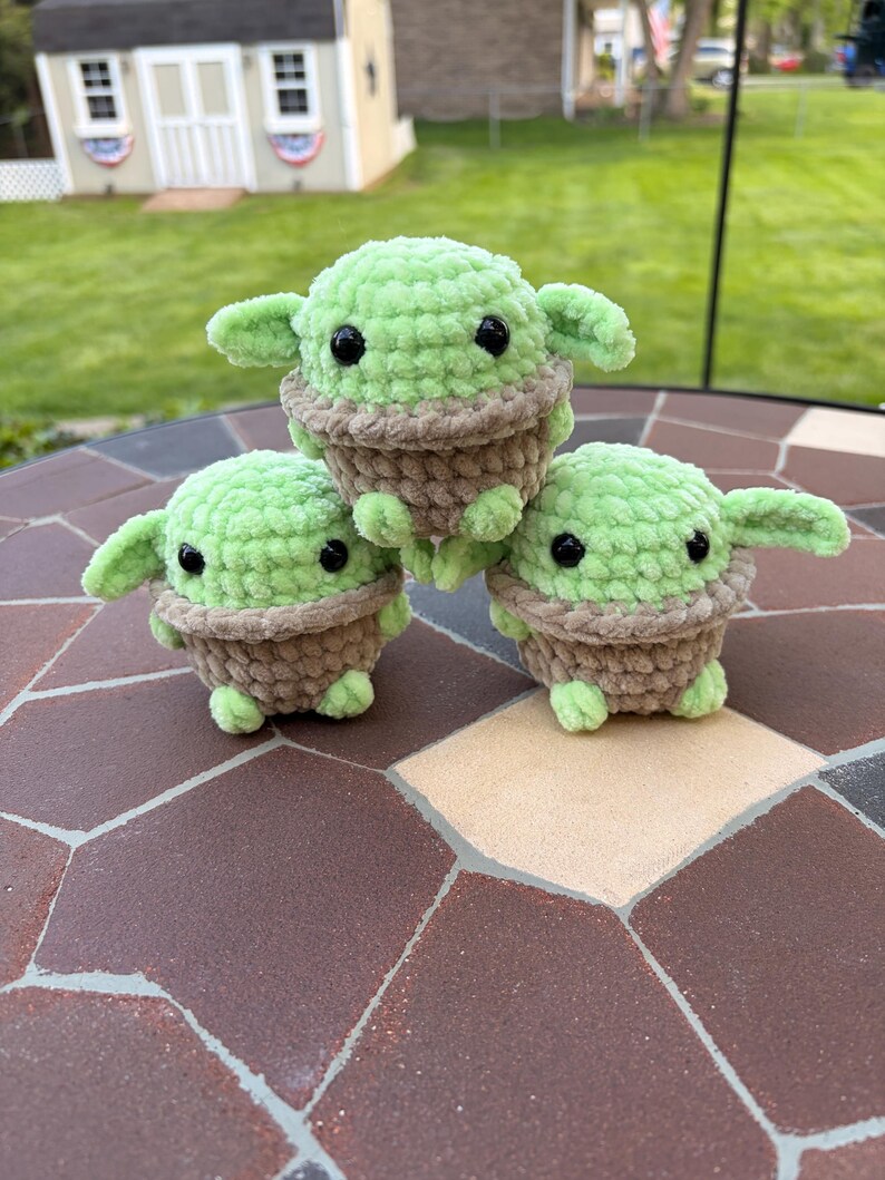 May include: Three handmade crochet figures of a character with large ears and black eyes. The figures are green with a brown base and are arranged on a brown and tan tiled surface. The figures are approximately 10 cm tall.