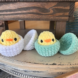 May include: Three crocheted chicks in eggs. One chick is in a white egg, one is in a green egg, and the third is in a green egg that is open. The chicks are yellow with black eyes and orange beaks.