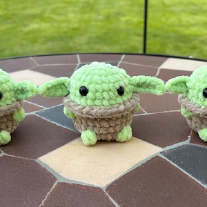 May include: Three handmade crochet figures resembling a popular character. Each figure has a light green head and ears, a brown body, and black eyes. The figures are displayed on a brown and tan tiled surface.