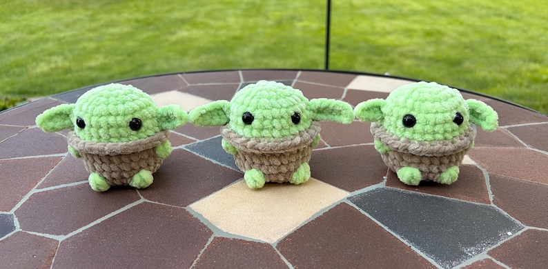 May include: Three handmade crochet figures resembling a popular character. Each figure has a light green head and ears, a brown body, and black eyes. The figures are displayed on a brown and tan tiled surface.