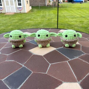 May include: Three green and brown crocheted figures with large ears and black eyes are displayed on a mosaic tile surface. The figures are arranged in a row, showcasing a handmade craft item.