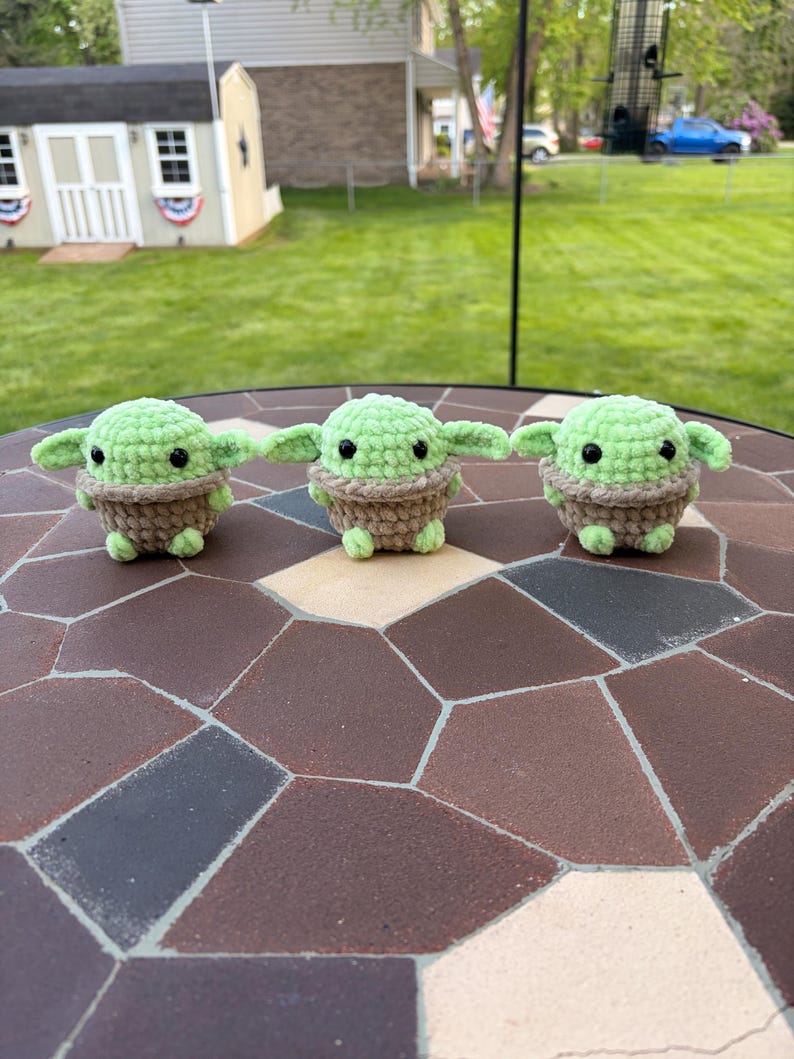 May include: Three green and brown crocheted figures with large ears and black eyes are displayed on a mosaic tile surface. The figures are arranged in a row, showcasing a handmade craft item.