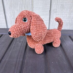Pode incluir: Um brinquedo de pelúcia de dachshund de crochê com pelo marrom e olhos azuis. O brinquedo está sentado em uma superfície de madeira.