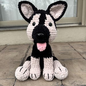 May include: A crocheted plush toy dog with black and white fur, green eyes, and a pink tongue. The dog is sitting on a tiled surface.