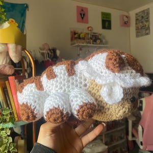 May include: A crocheted plush toy resembling a white and brown furry creature with a rounded body and four legs. The toy has a white face with brown eyes and a small, white, crocheted flower on its head.