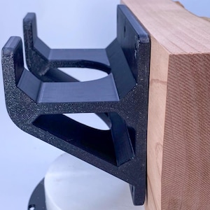 May include: A black, 3D-printed bracket with a textured surface, mounted to a wooden block. The bracket has a geometric design with open spaces and is designed to hold an item. The wood has a natural grain.