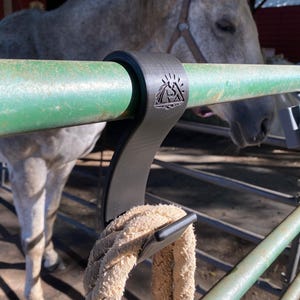 May include: A black metal hook designed to hang over a green metal fence rail. The hook has a mountain and sun logo. A beige rope is hanging from the hook. A horse is in the background.