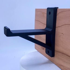 May include: A black metal shelf bracket attached to a light brown wooden board. The bracket has a right-angle design with a support beam. The bracket is designed to hold a shelf. The wood grain is visible.