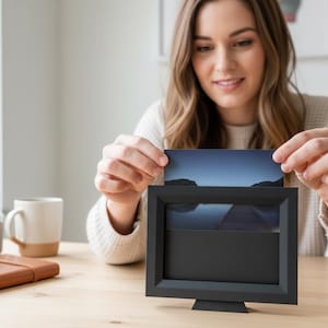 Puede incluir: Un marco de fotos negro con una imagen de paisaje que se está insertando. El marco es rectangular con un soporte. La imagen muestra una masa de agua y un cielo oscuro. Una mujer sostiene la foto.