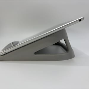 Puede incluir: Un soporte gris para tablet con un soporte triangular y una base curva. Una tableta plateada descansa sobre el soporte, en ángulo para su visualización. El soporte está hecho de un material liso y sólido.
