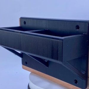 May include: A black, 3D-printed wall-mounted organizer with two compartments. The organizer is mounted on a wooden surface and features a bracket for support. The design includes screw holes for installation.
