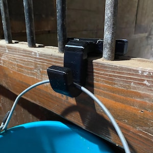 May include: A black plastic hook attached to a wooden beam and metal bars. A gray wire is attached to the hook, and a blue bucket is visible below. The hook is designed to hang items.