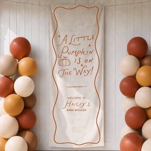 May include: A vertical off-white fabric banner with a wavy orange border and the text A LITTLE PUMPKIN IS ON THE WAY! WELCOME TO HAILEY'S BABY SHOWER in orange. Two balloon columns in shades of cream, tan, and burnt orange flank the banner.